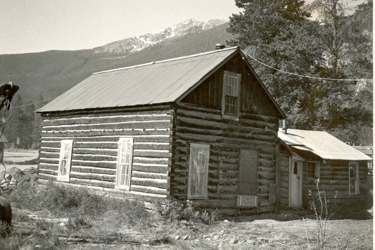 Black and white photo of Bailey House at its original location with snow covered mountains in the background.