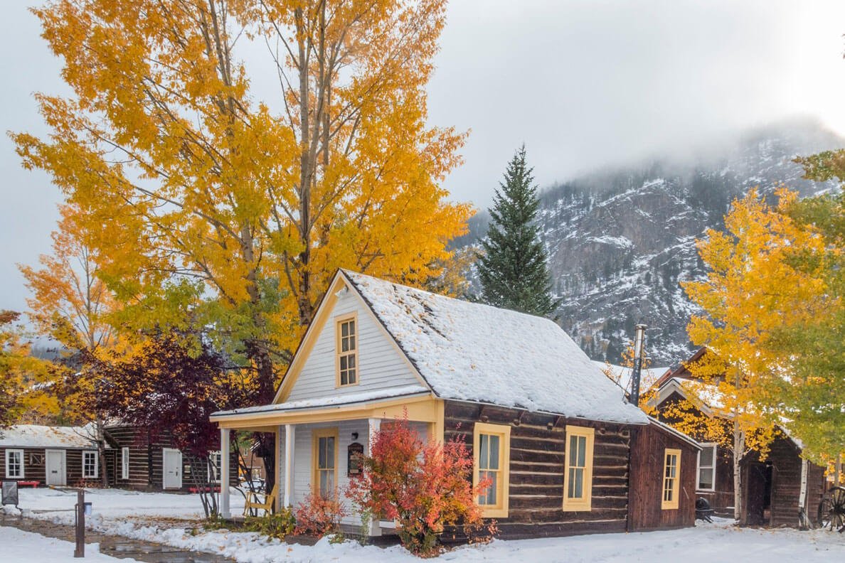 Snow covered cabins with golden aspens and snow falling on the mountains in the background