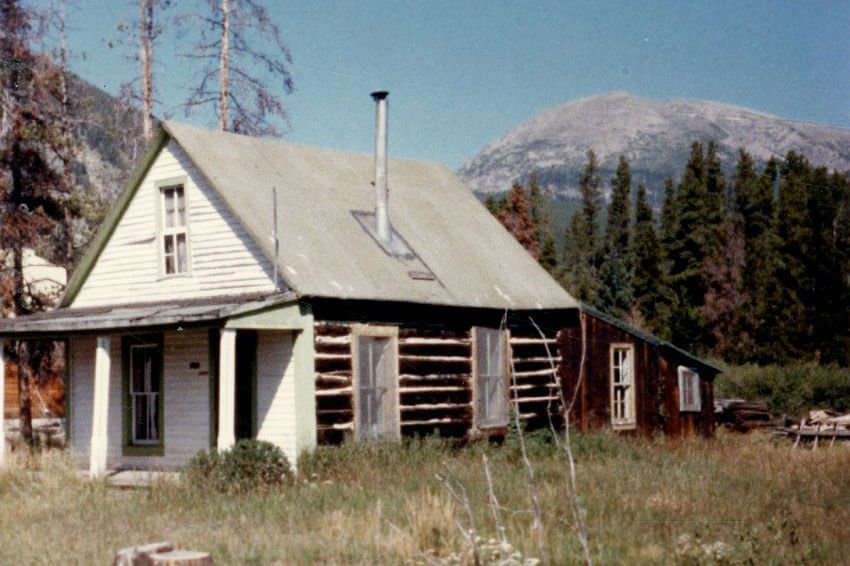 Old color photo of cabin in meadow with snow covered mountains in background