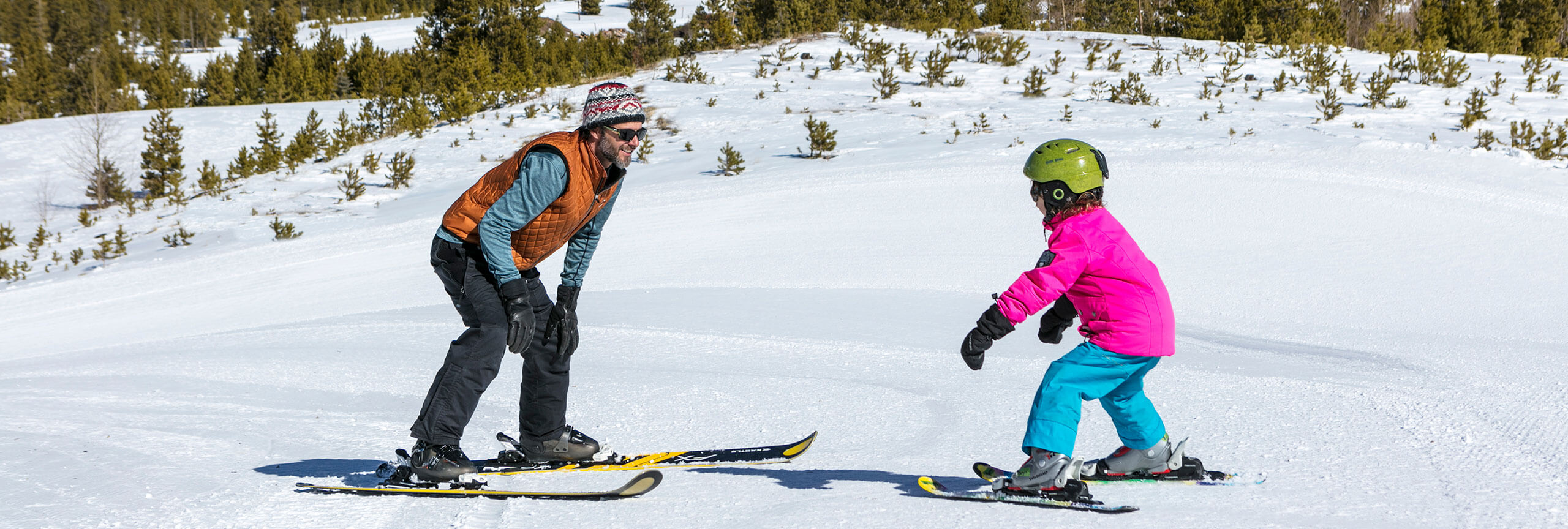 Man skiing backwards looking up hill at girl learning to ski