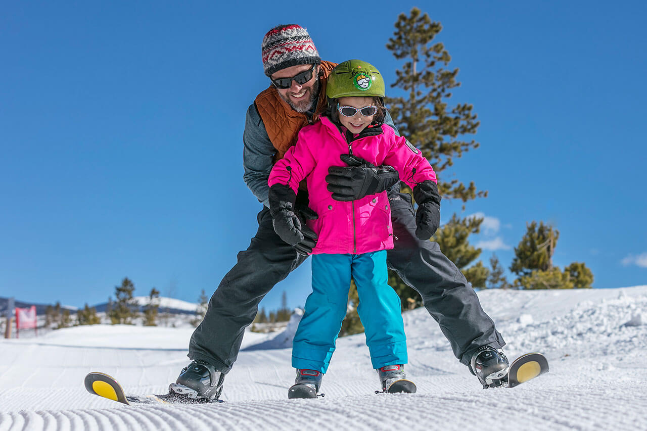 Man skiing with girl in front of him helping her learn to ski on beginner ski hill