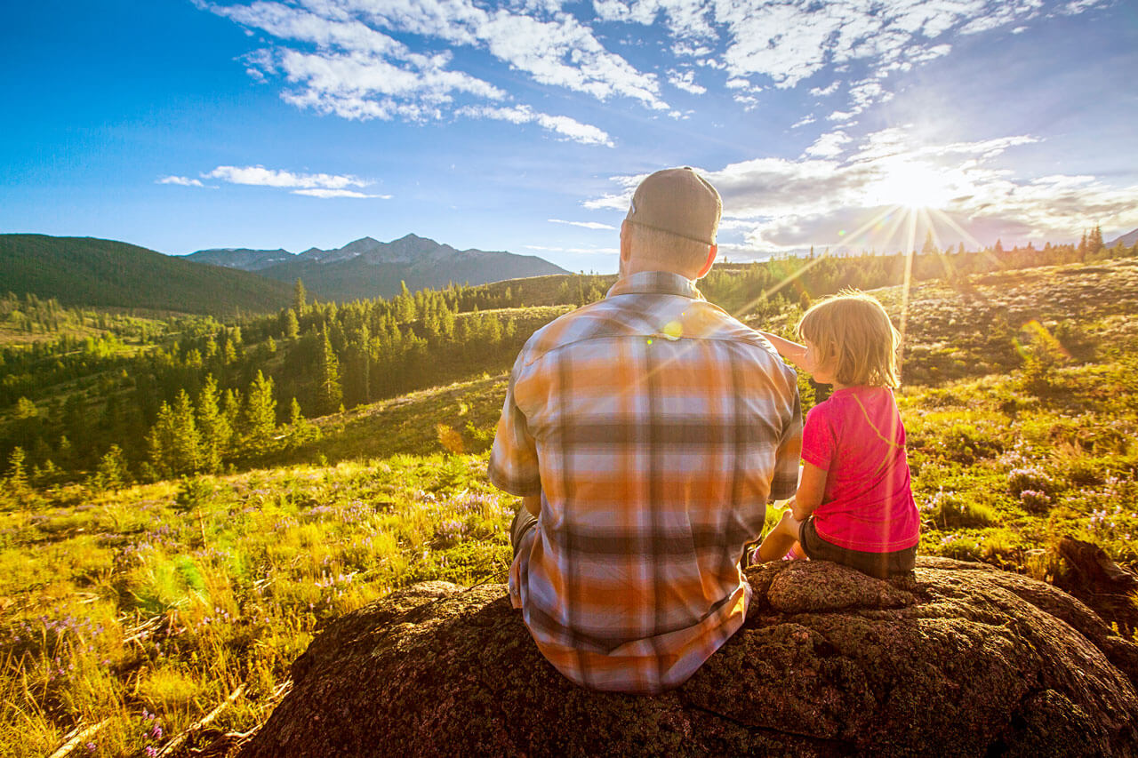 Father and child sitting on a rock looking out at mountains during sunset.
