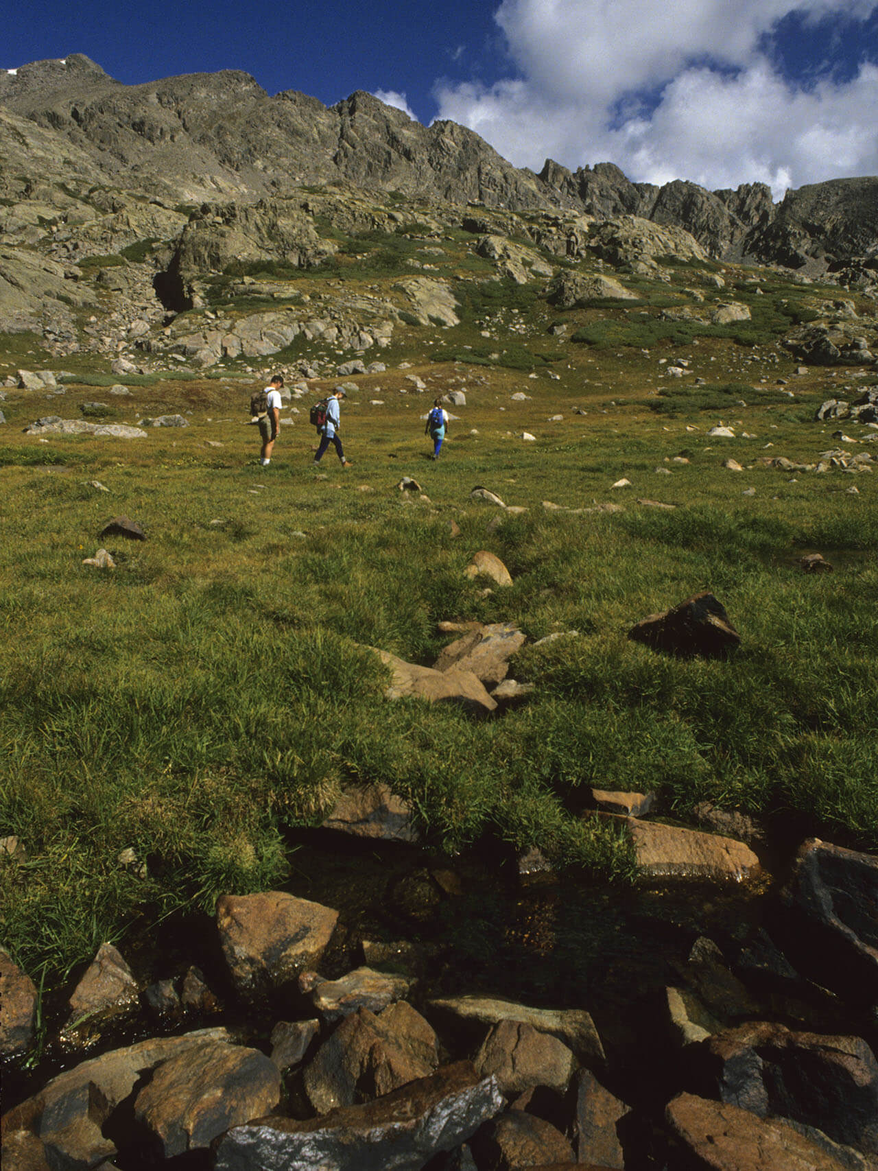 Three hikers on alpine tundra with craggy peaks in the background.