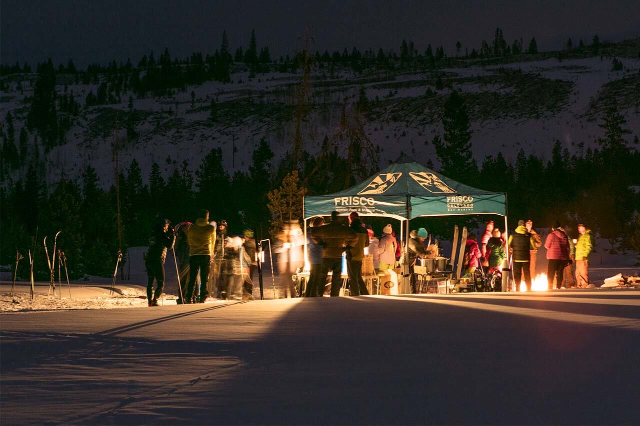 Group of people standing around tent and fire pit at Eat, Ski & Be Merry