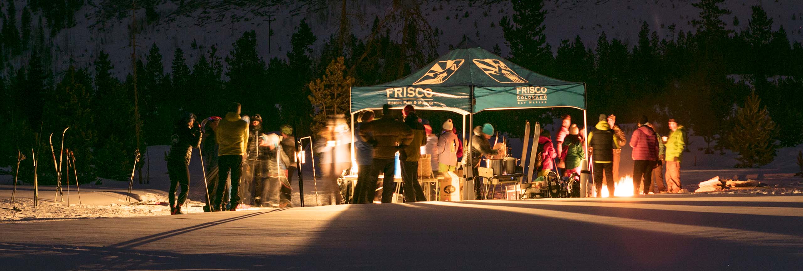 Group of people standing around tent and fire pit at Eat, Ski & Be Merry
